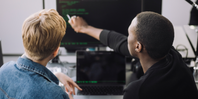 Color photo of Black man seated next to a white man and pointing to a desktop monitor displaying lines of code 